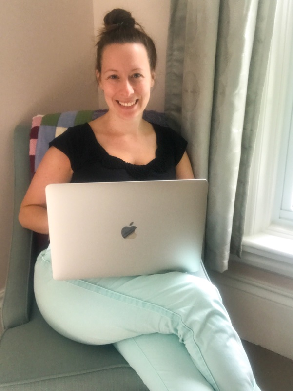 Jenn sitting in a chair with her laptop. She is wearing blue pamts and a black top